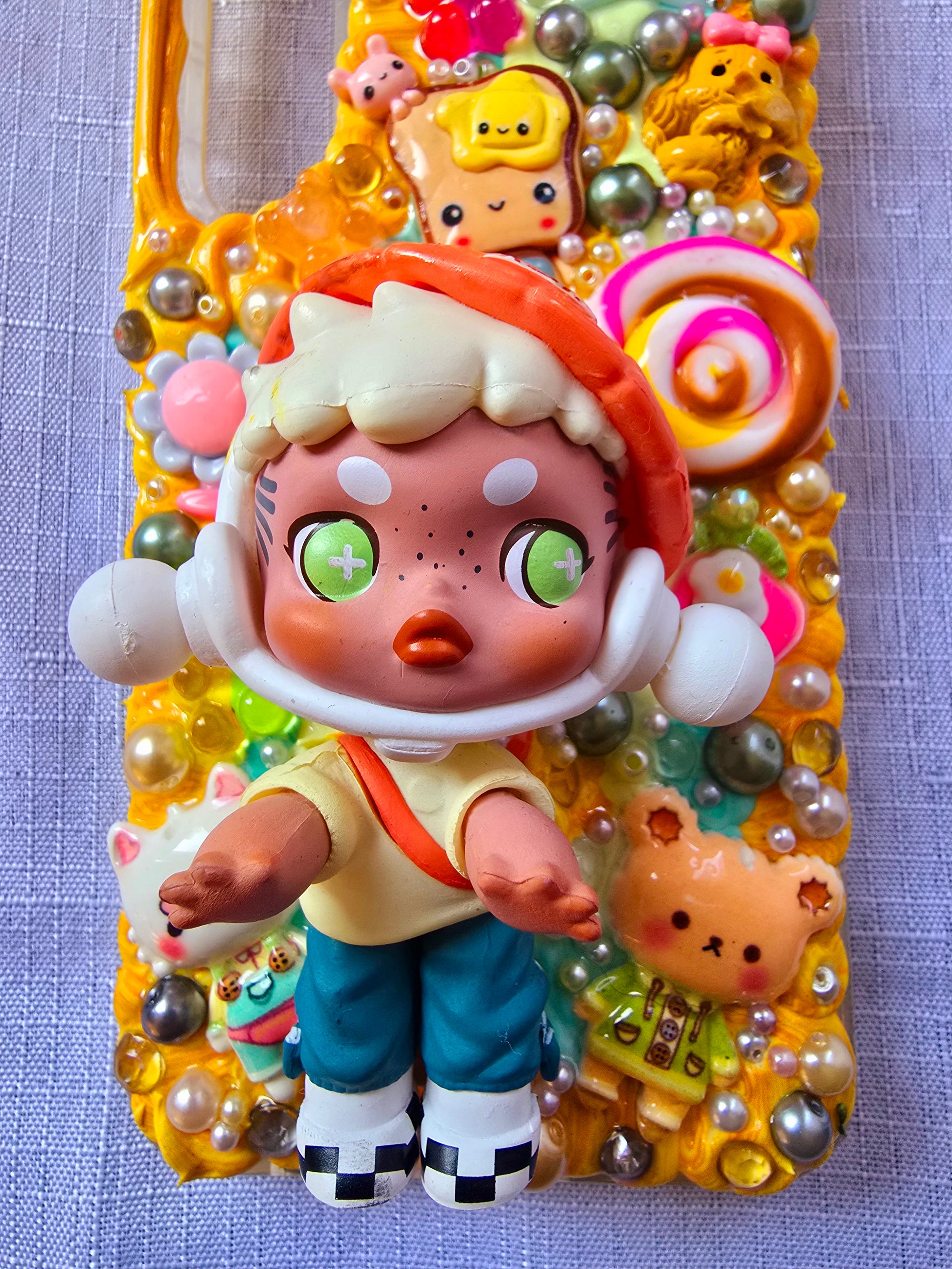 Handmade Samsung S23 Plus phone case with colorful kawaii and toy-themed 3D decorations, featuring a doll figure, pearls, cartoon charms, and candy accents on a yellow base.