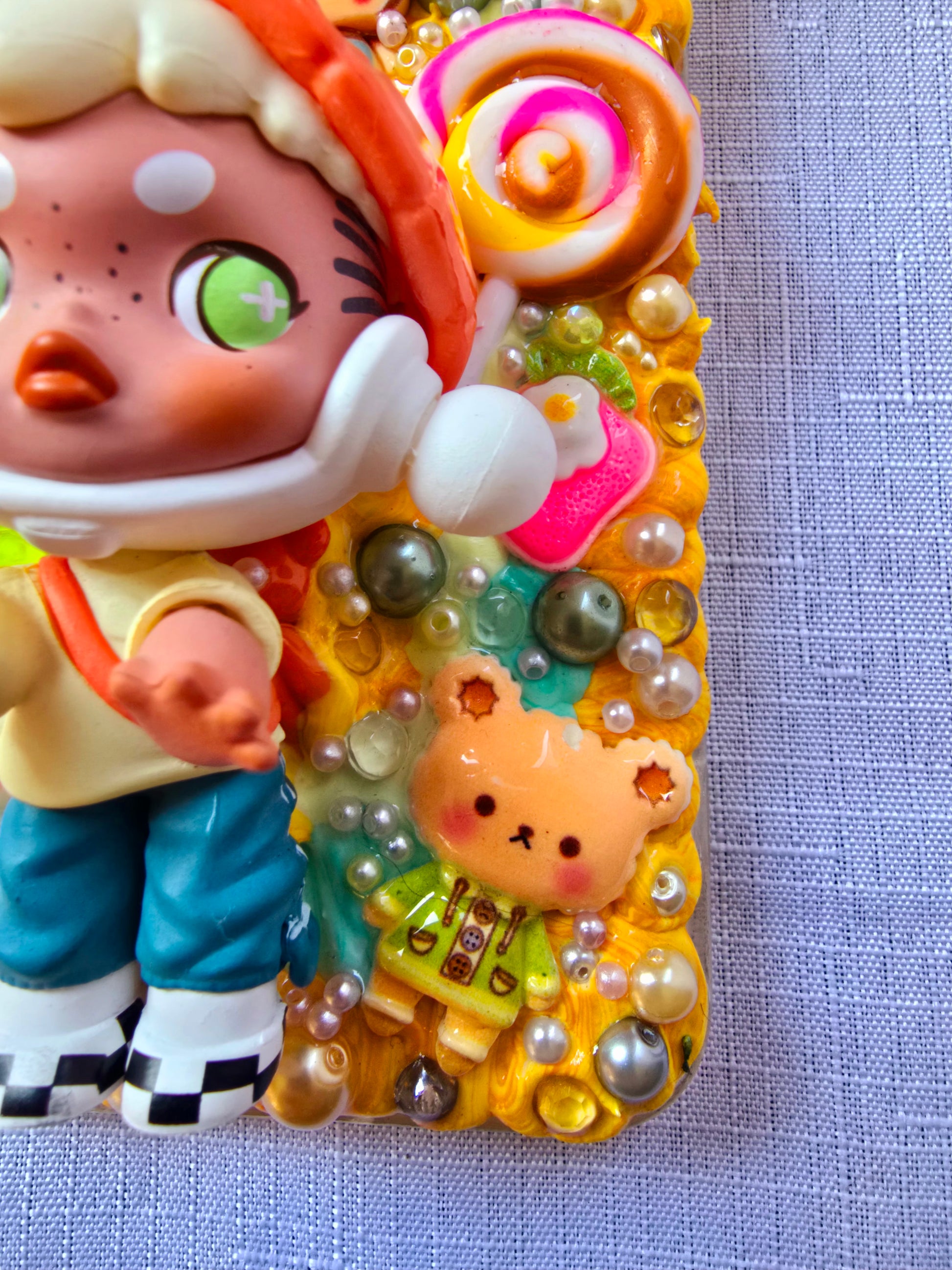 Handmade Samsung S23 Plus phone case with colorful kawaii and toy-themed 3D decorations, featuring a doll figure, pearls, cartoon charms, and candy accents on a yellow base.