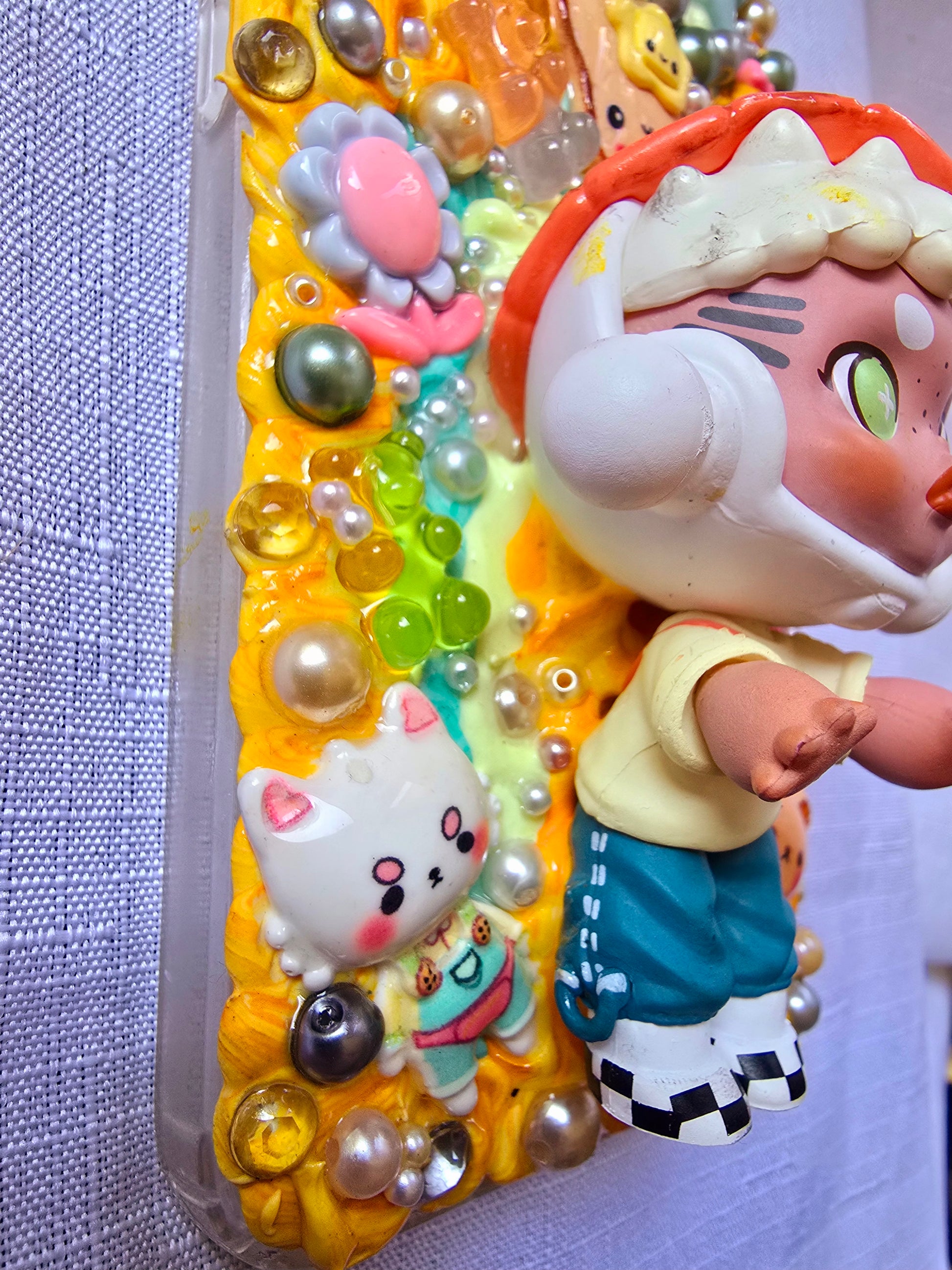 Handmade Samsung S23 Plus phone case with colorful kawaii and toy-themed 3D decorations, featuring a doll figure, pearls, cartoon charms, and candy accents on a yellow base.
