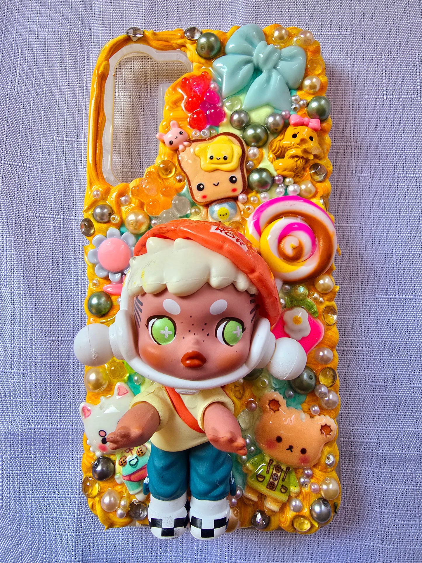Handmade Samsung S23 Plus phone case with colorful kawaii and toy-themed 3D decorations, featuring a doll figure, pearls, cartoon charms, and candy accents on a yellow base.